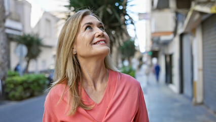 A smiling middle-aged blonde woman looks up as she strolls outdoors on a city street, exuding a...