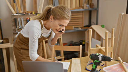 Blonde woman multitasking in a carpentry workshop while talking on a phone and writing notes.