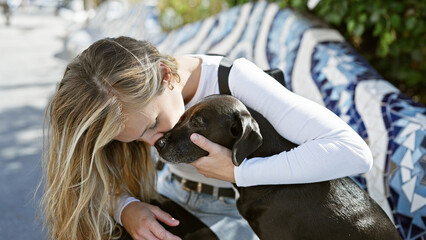 A young caucasian woman lovingly embraces her black labrador on a bench in a sunny outdoor park.