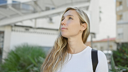 A young, beautiful, blonde woman standing thoughtfully on a city street, embodying urban lifestyle.