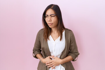 Middle age chinese woman wearing glasses over pink background with hand on stomach because nausea, painful disease feeling unwell. ache concept.