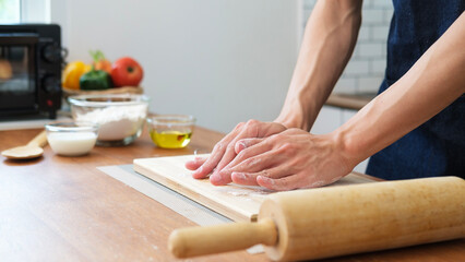 Unrecognizable young man using a rolling pin to roll over bread doughs on wooden table in kitchen.