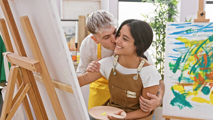 A man kisses a woman's cheek in an art studio, both holding paintbrushes and wearing aprons near...