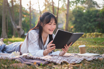 Carefree young Asian woman lying on picnic blanket and reading book.