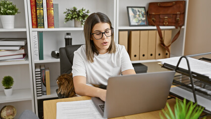Focused hispanic woman working on laptop in office with dog and bookshelf.