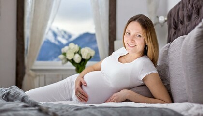 A young pregnant woman is lying on the bed wearing all wihite, smiling in the room