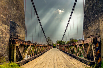 A woman walks across a historic suspension bridge toward a threatening, dramatic sky. Features...
