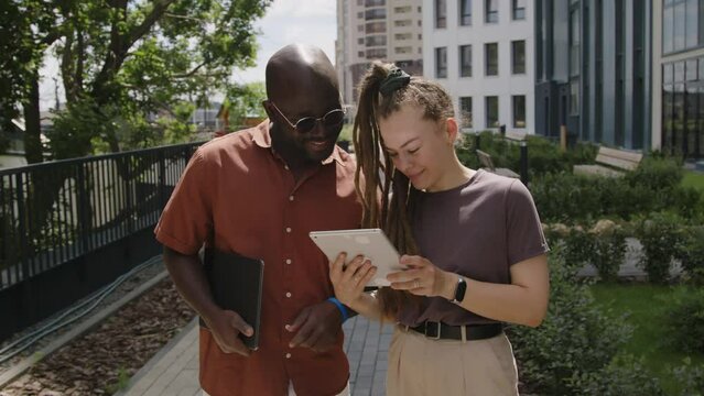 Medium footage of Caucasian businesswoman with dreadlocks presenting project ideas on tablet to cheerful African American associate during walk in urban park