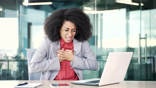 Sick young african american businesswoman suffering from chest tension, heart attack working on laptop sitting at desk at workplace in business office. Black woman needs urgent medical attention