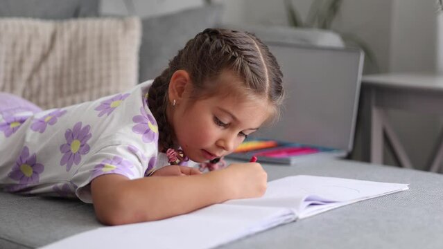 Side view of adorable little girl with beautiful braid hairstyle lying on sofa at home and drawing picture for mom