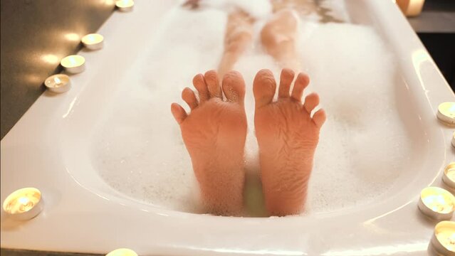 happy person taking bath lies in white soap foam, legs feet close-up fingers dancing joyfully, skin smacks in hot warm water, wet body part washing in white bathtub. Relax calm aroma candles light spa