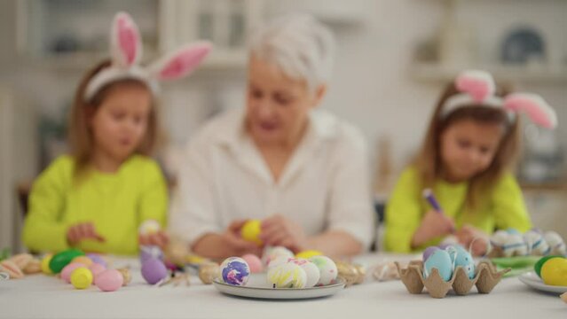 Easter grandmothers with granddaughters. Smiling grandmother with twins grandchildren painting decorating eggs in rabbit bunny ears, talking, celebrating together at home. Easter tradition concept.