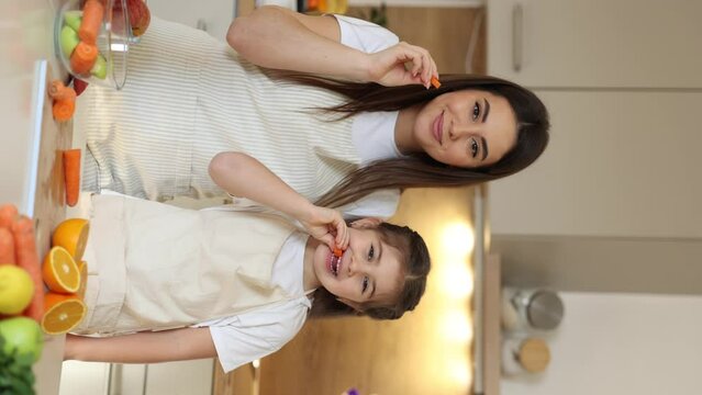 Young mom and cute daughter in aprons try carrots in the kitchen. Happy family sliced fruits and vegetables at home. Vertical video