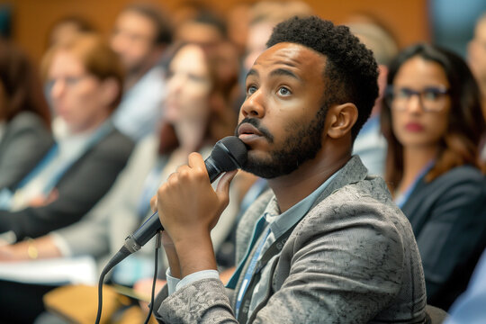 Multiethnic male technology conference attendee using microphone to ask a question regarding the demonstration of an innovative device while seated in the audience