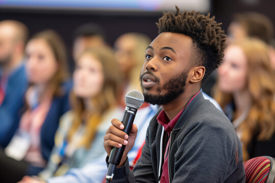 Multiethnic male technology conference attendee using microphone to ask a question regarding the demonstration of an innovative device while seated in the audience