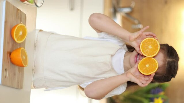 Adorable little girl in apron stand in kitchen and smiling. Child holding halves of oranges at her eyes. Vertical video