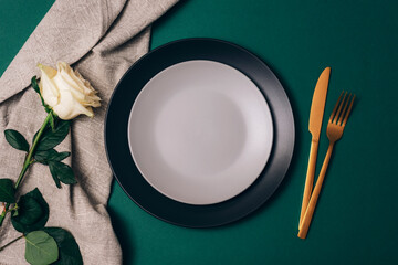 Table place setting with white rose flower, plates and golden cutlery on dark green background. Top...