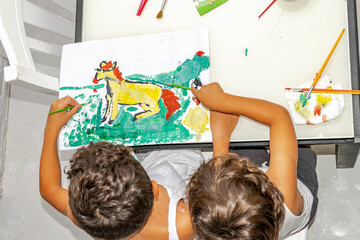 Two unrecognisable little boys sitting at home, painting a horse in a meadow with a brush on a...
