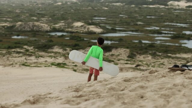 Boy stops on dunes to look out into distance wetlands while sand boarding - wide shot