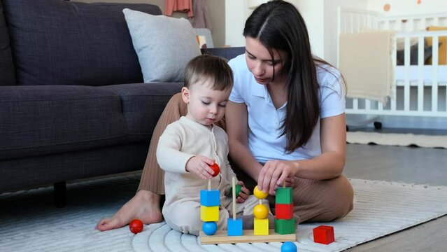 Caucasian mother and little son playing with wooden toys on floor in living room. Construction development using toy bricks, spending leisure time together.