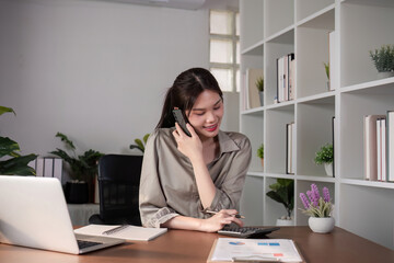 Young Asian business woman sits on the phone in an online business meeting using a laptop in a modern home office decorated with shady green plants.