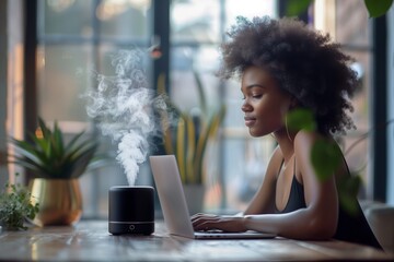 black essential oil diffuser on the table in the office, African girl is sitting with a laptop. Concept aromatherapy and relaxing. Air freshener - obrazy, fototapety, plakaty