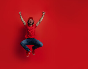 20s white handsome man in a red t-shirt and blue jeans jumping in the air with arms raised, smiling and celebrating something on red background 