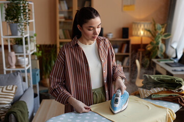 High angle portrait of young woman ironing clothes on laundry day and taking care of house chores copy space