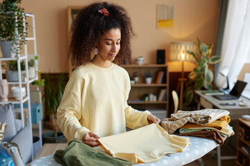 Waist up portrait of smiling young woman with big curly hair folding clothes on ironing board at home copy space