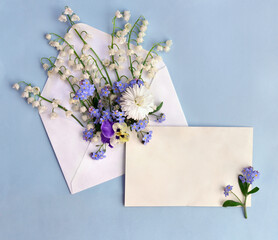 White flowers Lily of the valley ( Convallaria, May bells, may lily ), viola, forget-me-not, daisy, postal envelope with paper card note with space for text on a blue background. Top view, flat lay