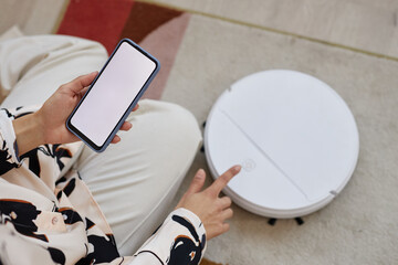 Top view closeup of unrecognizable young woman operating smart robot vacuum cleaner and holding...