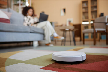 Close up of white robot vacuum cleaner on colorful carpet with blurred young woman in background...