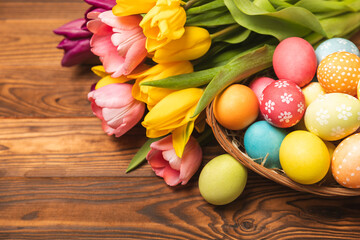 Fototapeta na wymiar Easter basket filled with colorful eggs and a bouquet of tulips on a textured wooden table. Easter celebration concept. Colorful easter handmade decorated Easter eggs. Place for text. Copy space