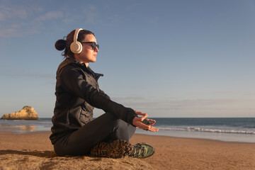 Sporty woman sitting meditating by the sea, wearing headphones and sunglasses.