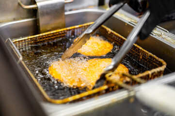 Close-up view of two golden-brown breaded fillets frying in bubbling oil, turned by tongs in a...