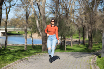 Fototapeta premium Young and beautiful brunette woman dressed in casual clothes and sunglasses, walks along a path between deciduous trees in autumn, in the background a lake.