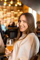 Young cheerful brunette woman looking at you while toasting with glass of beer