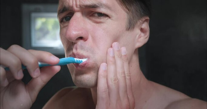 Close-up of man brushing his teeth in front of mirror in bathroom. Feeling of severe pain in tooth and writhing. Man holds his cheek with bad tooth with hand. Acute toothache does not go away.