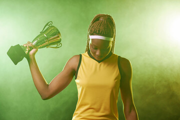 Waist up portrait of Black female athlete holding trophy cup and looking down with green smoke in...