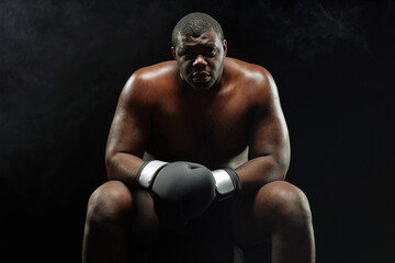 Fototapeta na wymiar Dramatic backlit portrait of muscular African American boxer looking at camera sitting against black background