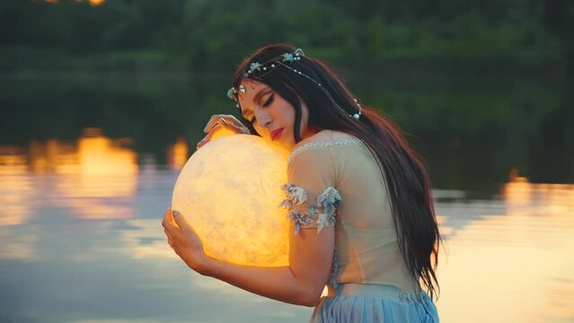 Fantasy woman embracing holding moon magic light glowing planet in hands hugs. Girl night elf beauty face fairy river nymph blue dress sitting in water dark sky lake. Lady queen art fortune teller 4k