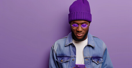 A man wearing a purple beanie and glasses is holding a cell phone. He is looking at the camera with a serious expression. man with phone on purple background - obrazy, fototapety, plakaty