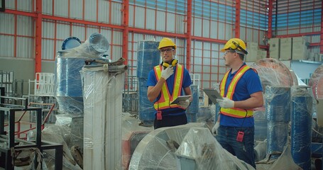 Industrial workers with tablet and headset in warehouse, evaluate machinery wrapped in protective plastic in an industrial warehouse during a quality check.