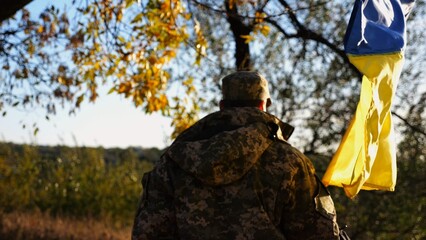 Young male military in uniform walks with waving flag of Ukraine at countryside. Soldier of ukrainian army going with blue-yellow banner outdoor. Concept of victory against russian aggression. - 756617779