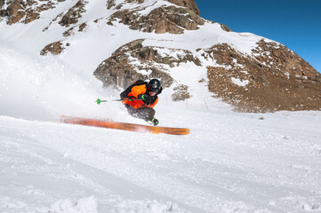 A skier in an orange jacket and a black helmet with a mask rides down a snowy slope at high speed....