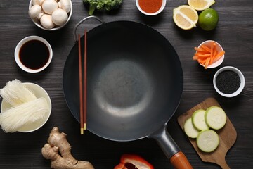 Empty iron wok and chopsticks surrounded by ingredients on dark grey wooden table, flat lay