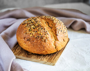 Freshly baked wheat bread topped with sesame and pumpkin seeds,  whole loaf lying on wooden cutting board, on white table cloth with kitchen towel beside. Homemade food concept