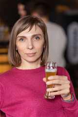 Young cheerful dark blond woman looking at you while toasting with glass of beer