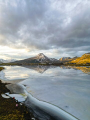 Tranquil landscape with icy lake in the foreground and snow-capped mountains under cloudy skies.