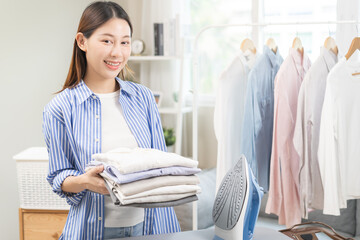 Happy smile asian young woman holding stack, pile folded hygiene laundry clothes at ironing board...